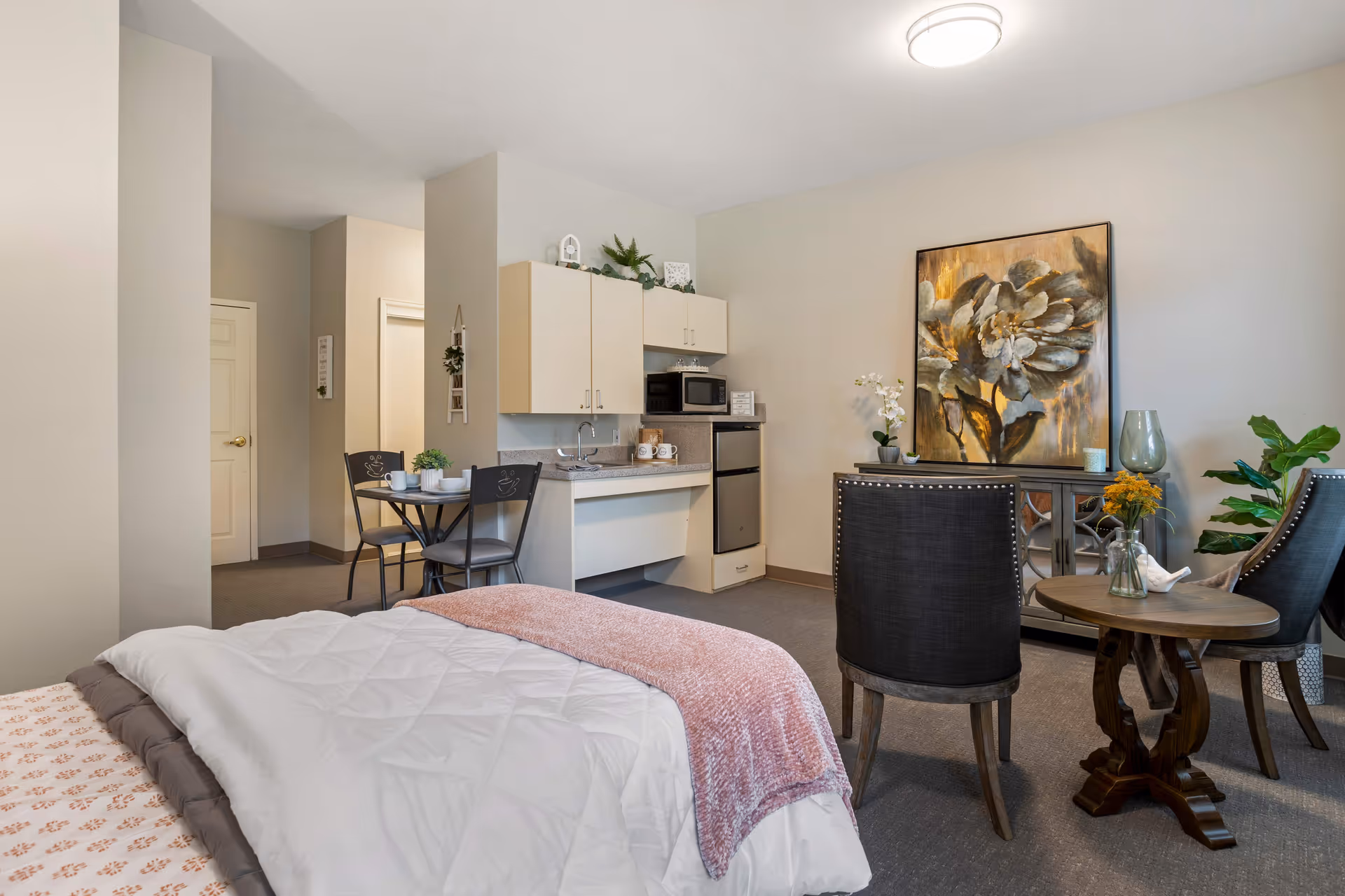 A senior living facility room with a bed covered in white and pink bedding in the foreground. The room includes a small kitchenette with a sink, microwave, and mini fridge. There is a small dining table with two chairs near the kitchenette. On the right side, there are two upholstered chairs and a small round wooden table with a vase of flowers. A large floral painting hangs on the wall above a cabinet, and the room is decorated with plants and soft lighting.