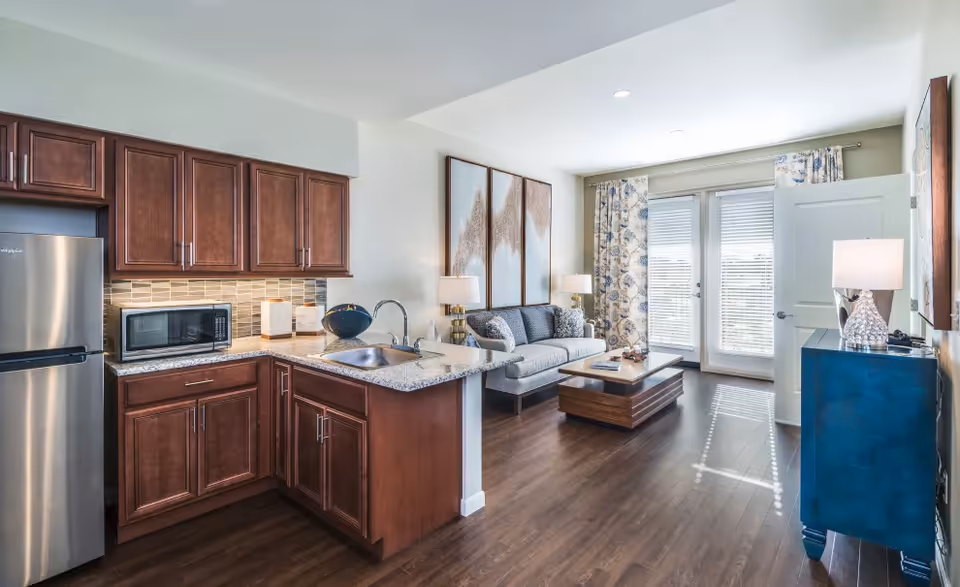 A bright and modern open-concept living space featuring a kitchen with dark wood cabinets, a stainless steel refrigerator, a microwave, and a granite countertop with a sink. Adjacent to the kitchen is a living room area with a gray sofa, a wooden coffee table, two table lamps, floral curtains, and glass doors letting in natural light.