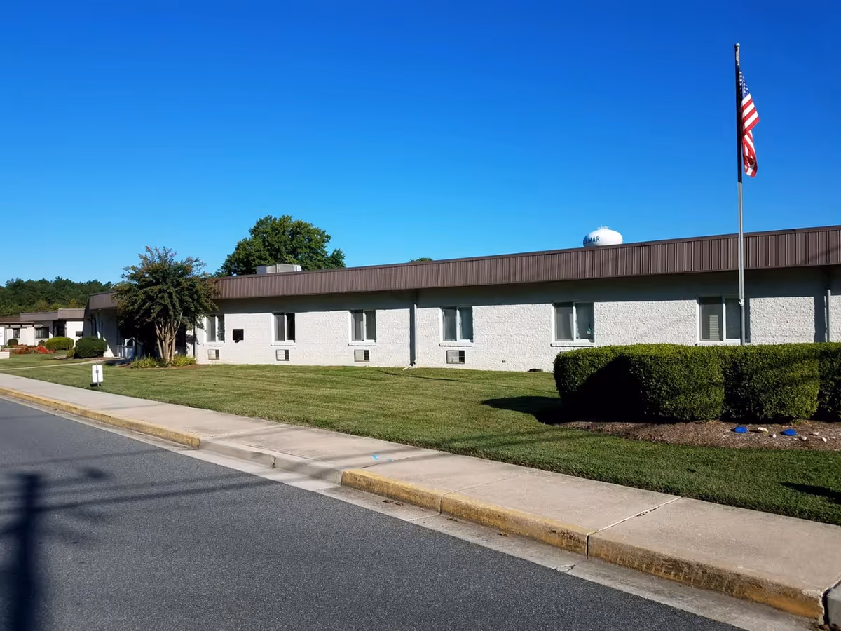 Single-story white brick nursing facility with a manicured lawn, an American flag on a pole, and a clear blue sky.