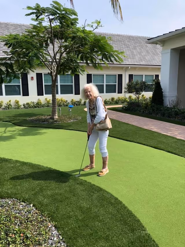 An elderly woman playing mini golf on a putting green outside at Courtyard Gardens Senior Living. She is holding a golf club and standing on a green artificial turf area with a building and landscaping in the background.