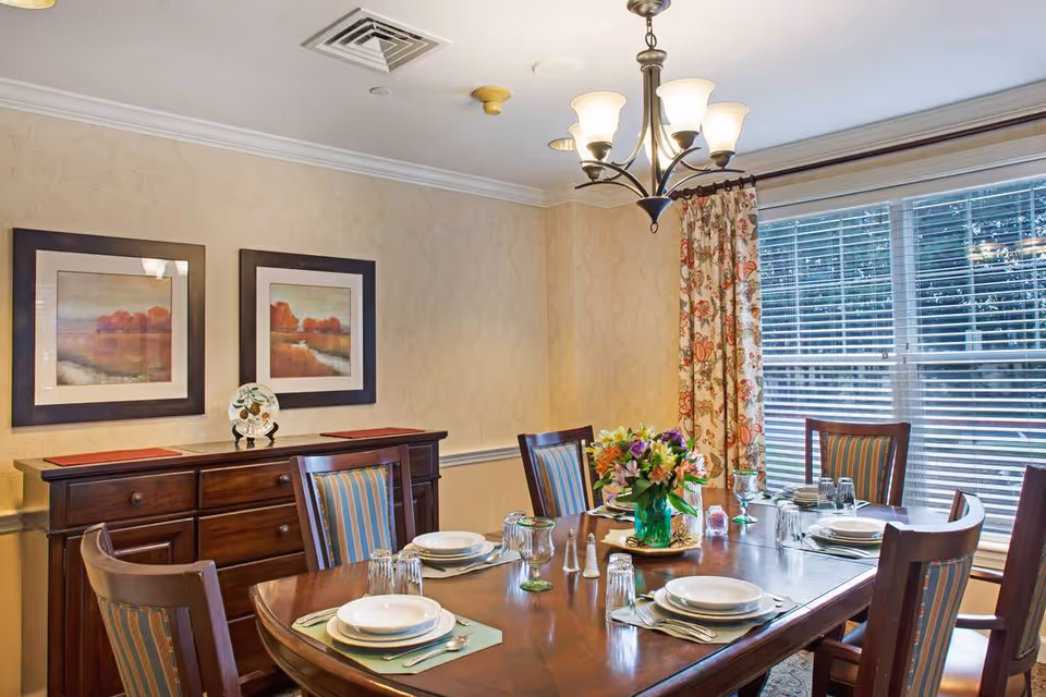 A dining room with a wooden table set for six people with plates, glasses, and silverware. A floral centerpiece is placed in the middle of the table. The room has a sideboard with two framed landscape paintings above it, floral curtains on the window, and a chandelier hanging from the ceiling.