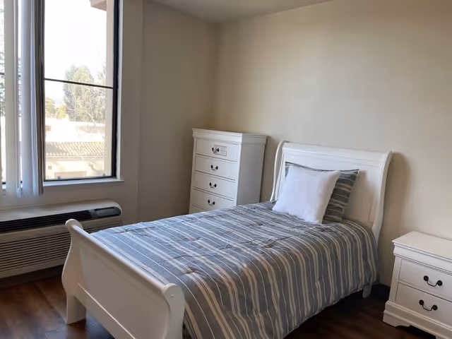 Single bedroom with a white sleigh bed, striped bedding, a tall dresser and nightstand next to a window and wall-mounted AC unit.