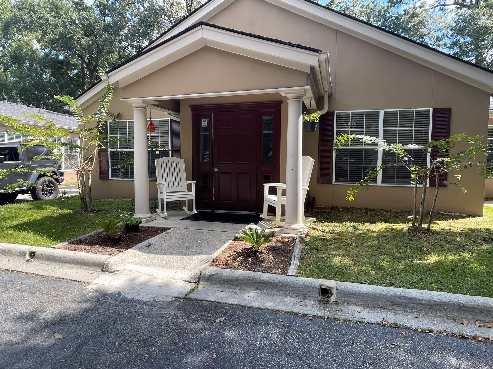 Front entrance of a beige building with a small covered porch supported by two white columns. Two white rocking chairs are placed on either side of a dark red door. There are windows with white blinds and dark red shutters on either side of the door. Small plants and shrubs are planted in mulched beds along the walkway leading to the entrance. A black vehicle is partially visible on the left side of the image, and trees are in the background.