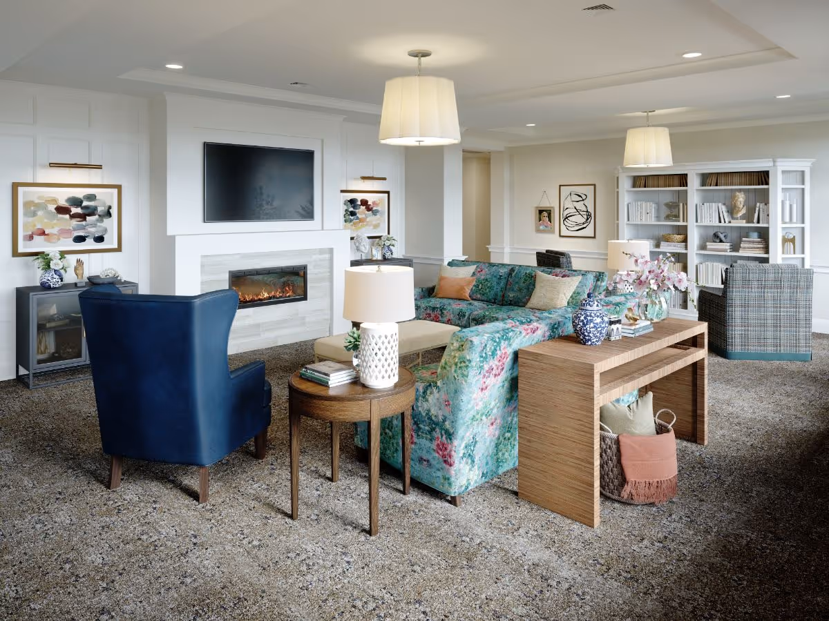 A cozy living room in a senior living facility featuring a floral patterned sectional sofa, a blue armchair, a wooden side table with a lamp and books, a modern electric fireplace beneath a mounted flat-screen TV, and a white bookshelf filled with books and decorative items. The room is carpeted and softly lit with ceiling lights and hanging lamps.