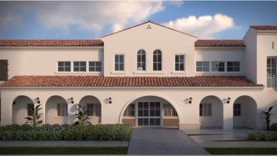 Front exterior view of a two-story building with white walls and a red tile roof, featuring arched doorways and windows, surrounded by greenery and a paved walkway leading to the entrance.
