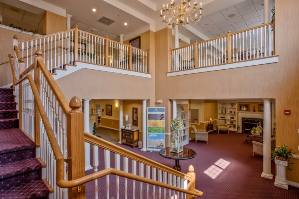 Two-story interior lobby with a carpeted staircase, balcony railings, chandelier, seating areas and decorative columns.