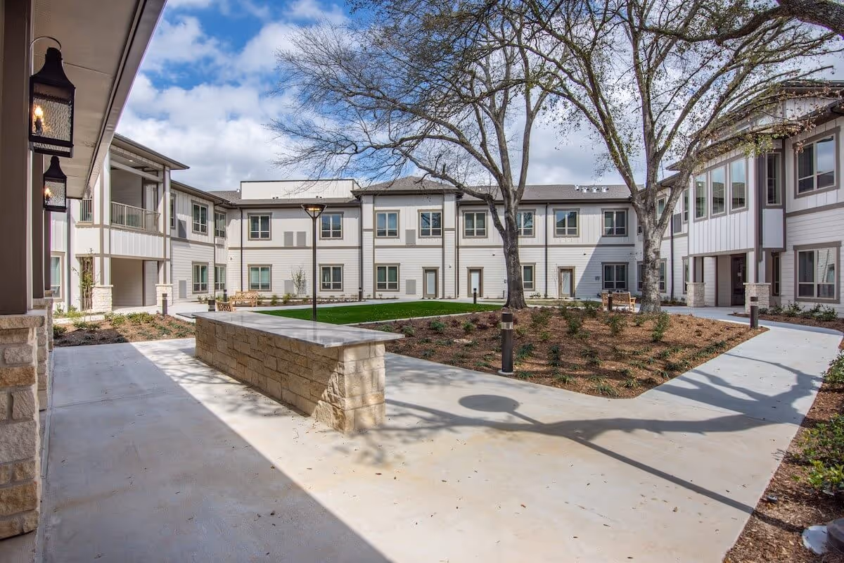 Sunny courtyard with paved walkways, landscaped beds, mature trees, and surrounding two-story white senior living buildings.