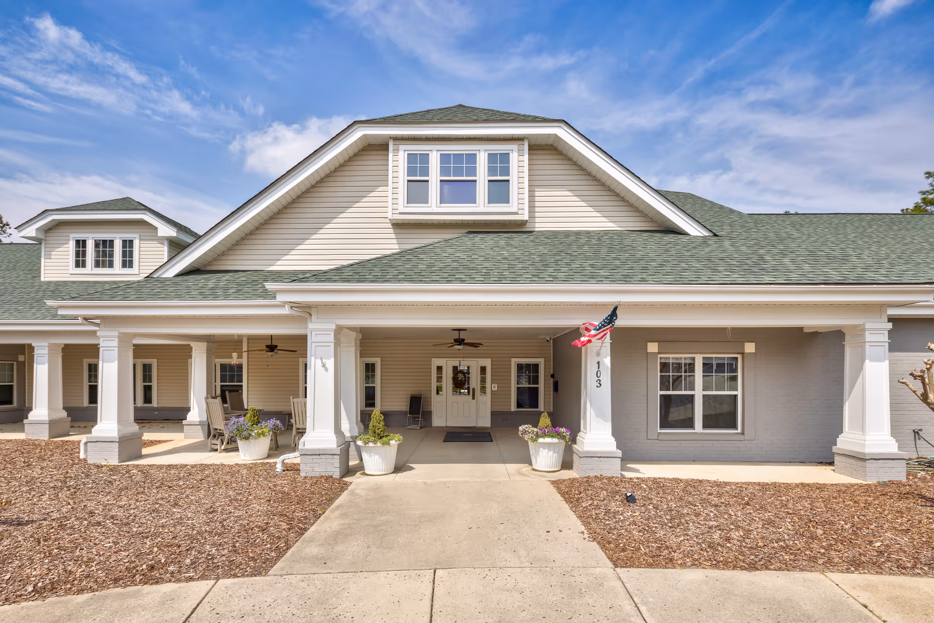 Front exterior view of a single-story building with a covered porch supported by white columns. The building has beige siding, green shingled roof, and several windows. There are potted plants and an American flag near the entrance with the number 103 displayed on a column.