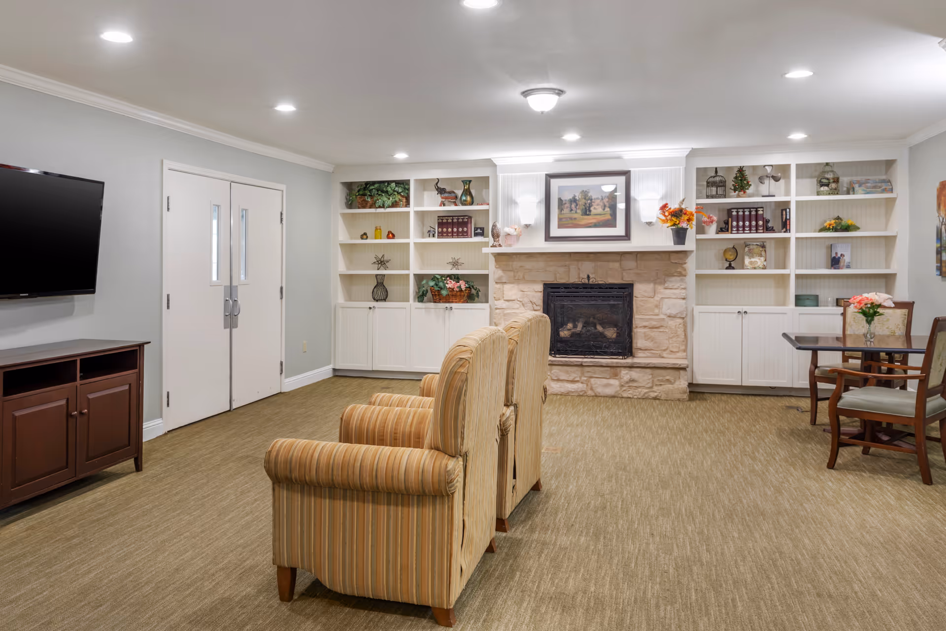 A cozy living room area with two striped armchairs facing a stone fireplace. The fireplace is flanked by white built-in shelves decorated with books, plants, and ornaments. A flat-screen TV is mounted on the wall to the left, above a dark wooden cabinet. To the right, there is a small table with chairs and a vase of flowers. The room has beige carpet and soft lighting from ceiling fixtures.