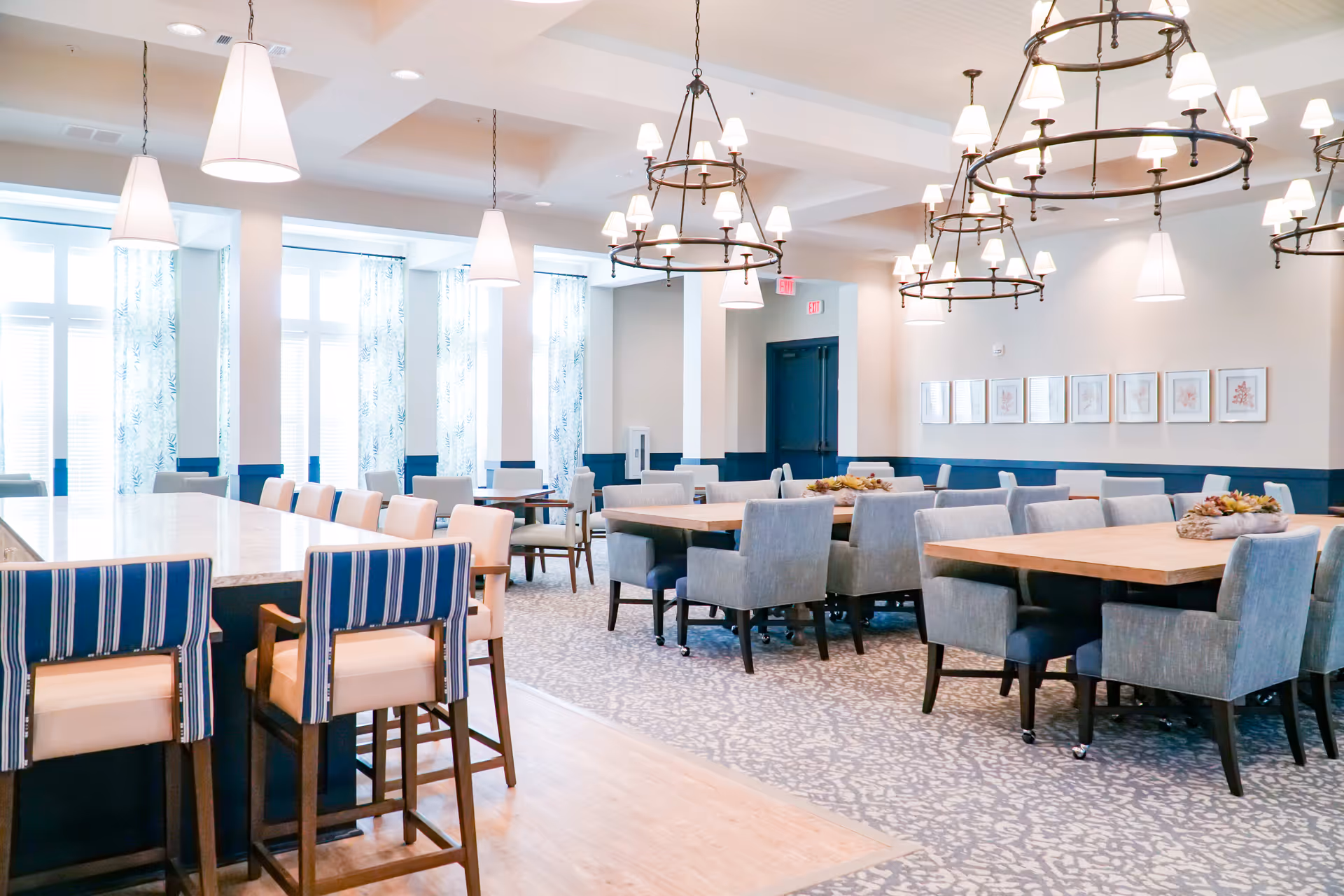 Bright and spacious dining room with multiple tables and chairs arranged neatly. The room features large windows with patterned curtains allowing natural light to fill the space. There are modern pendant lights and circular chandeliers hanging from the ceiling. The floor has a combination of wood and patterned carpet, and framed artwork is displayed on the wall.