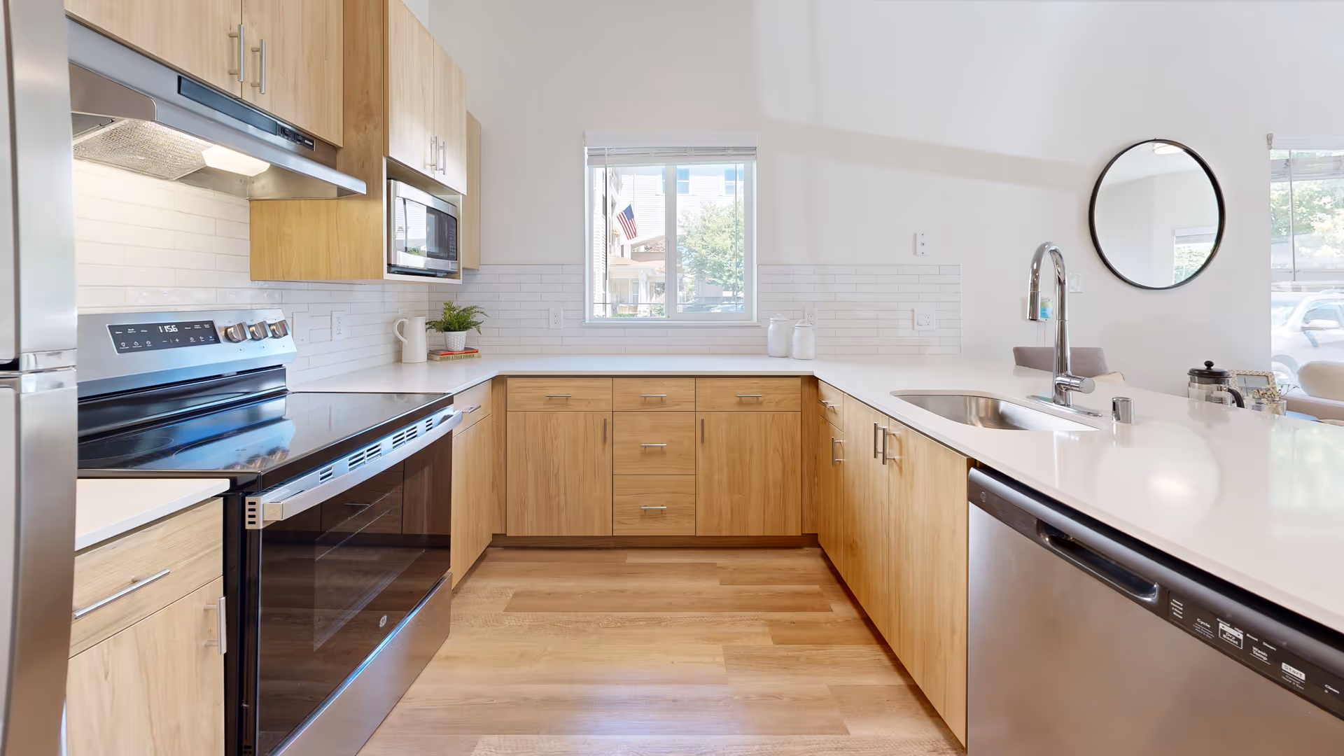 Bright modern kitchen with light wood cabinets, white countertops, stainless steel appliances including a stove, microwave, dishwasher, and refrigerator. A window above the counter lets in natural light, and a round mirror is mounted on the wall near the sink.