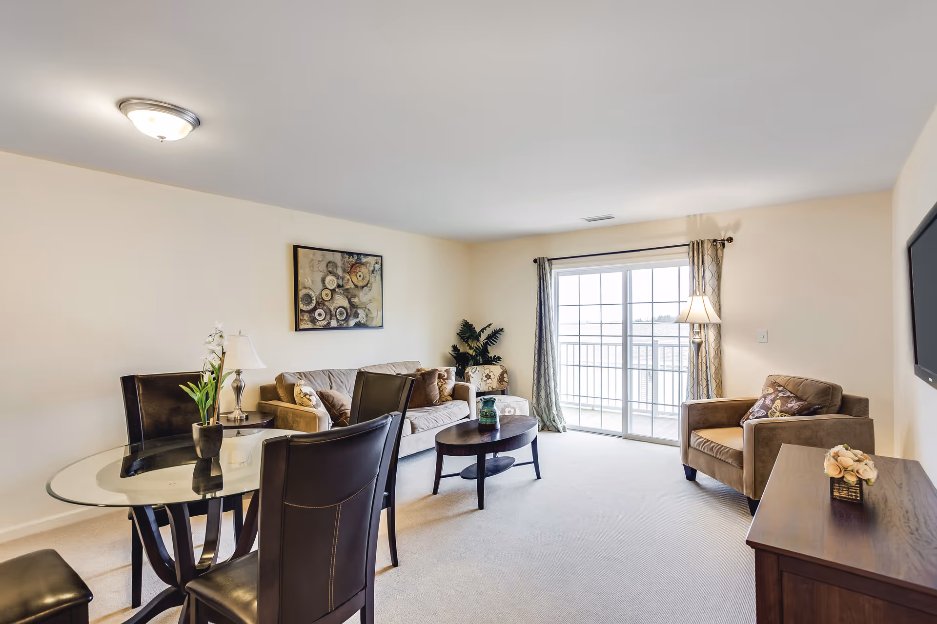 Bright living room with a sofa, armchair, glass dining table, coffee table and sliding glass doors to a balcony.