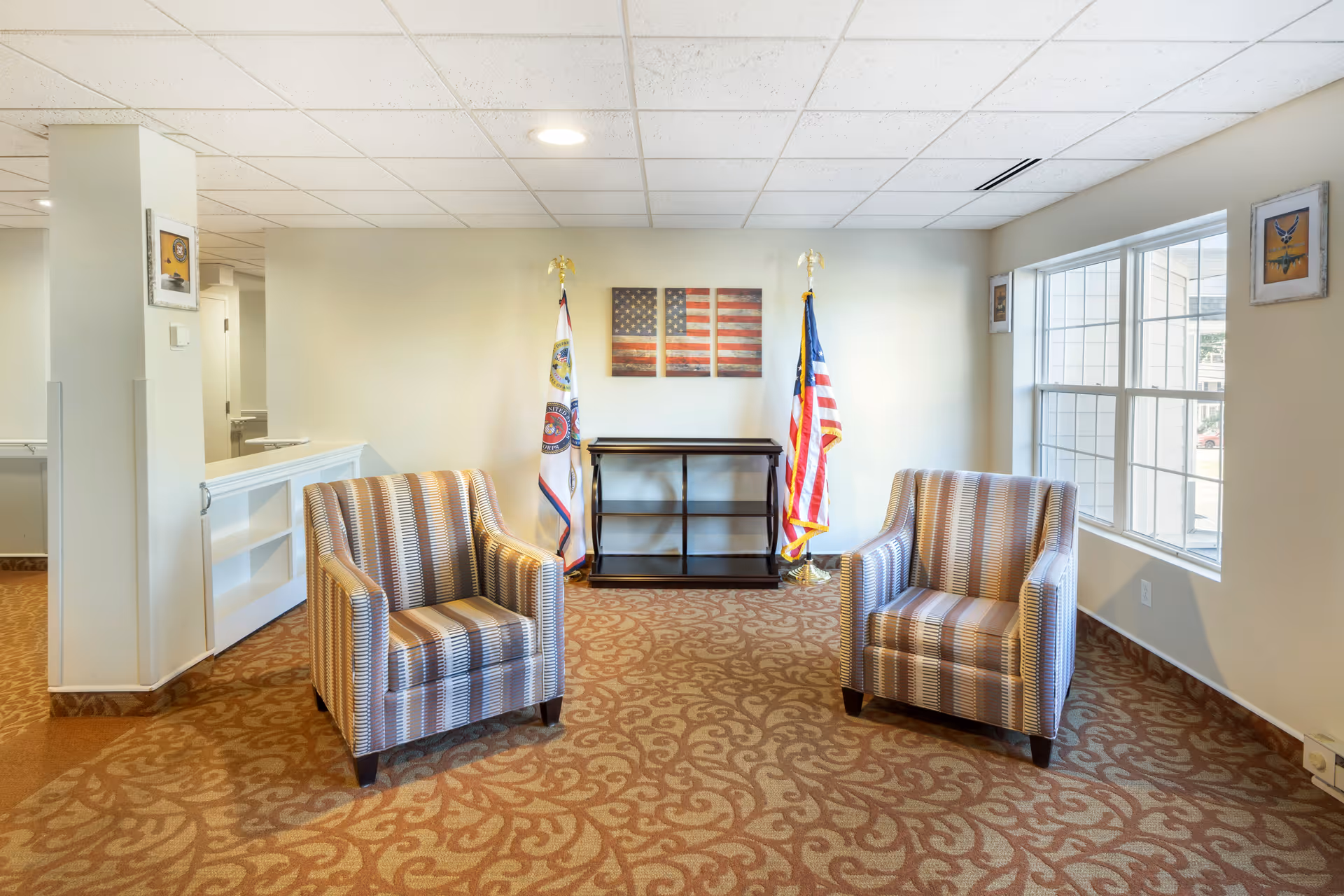 A bright sitting area with two striped armchairs, a small console table between two flags, and a large window on the right.