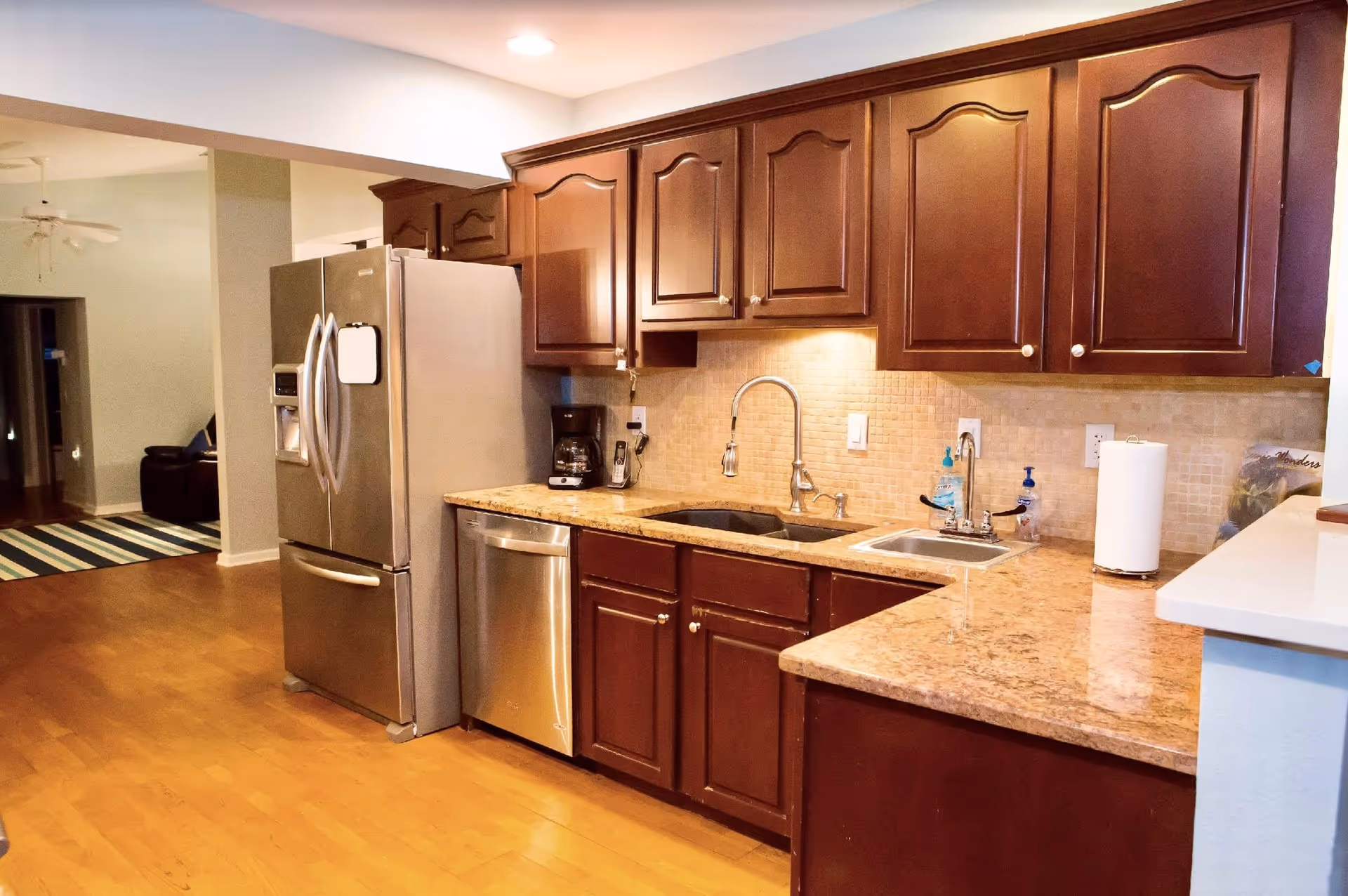 Interior view of a kitchen with wooden cabinets, granite countertops, a stainless steel refrigerator, dishwasher, and two sinks. The kitchen opens into a living area with a ceiling fan and a striped rug on the wooden floor.