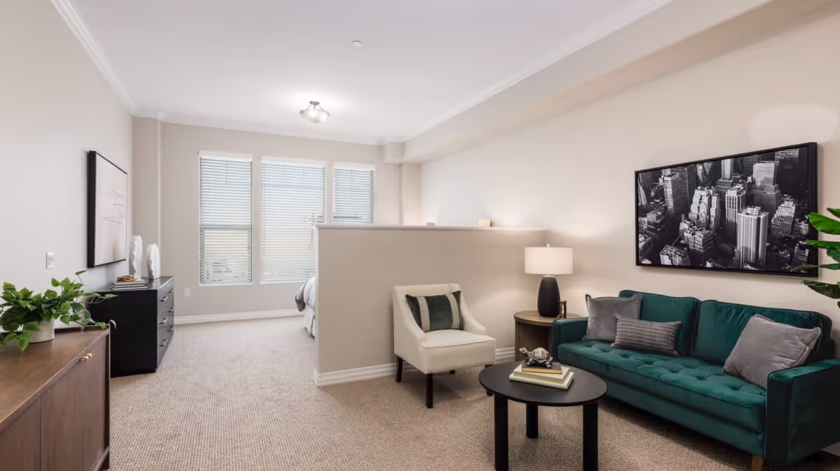A modern living area with a green sofa, a white armchair, a round black coffee table with books and a decorative item, a side table with a lamp, and a large black and white cityscape photo on the wall. Behind a half wall, a bedroom area with a bed is visible near three large windows with blinds. The room has beige walls and carpeted flooring.