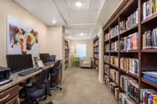 Interior view of a library or study area in a senior living facility. On the left side, there are desks with computers and chairs, and on the right side, there are tall bookshelves filled with books. At the end of the room, there is a comfortable armchair near a window with blinds. The walls are light-colored, and there is a colorful horse painting above the desks.