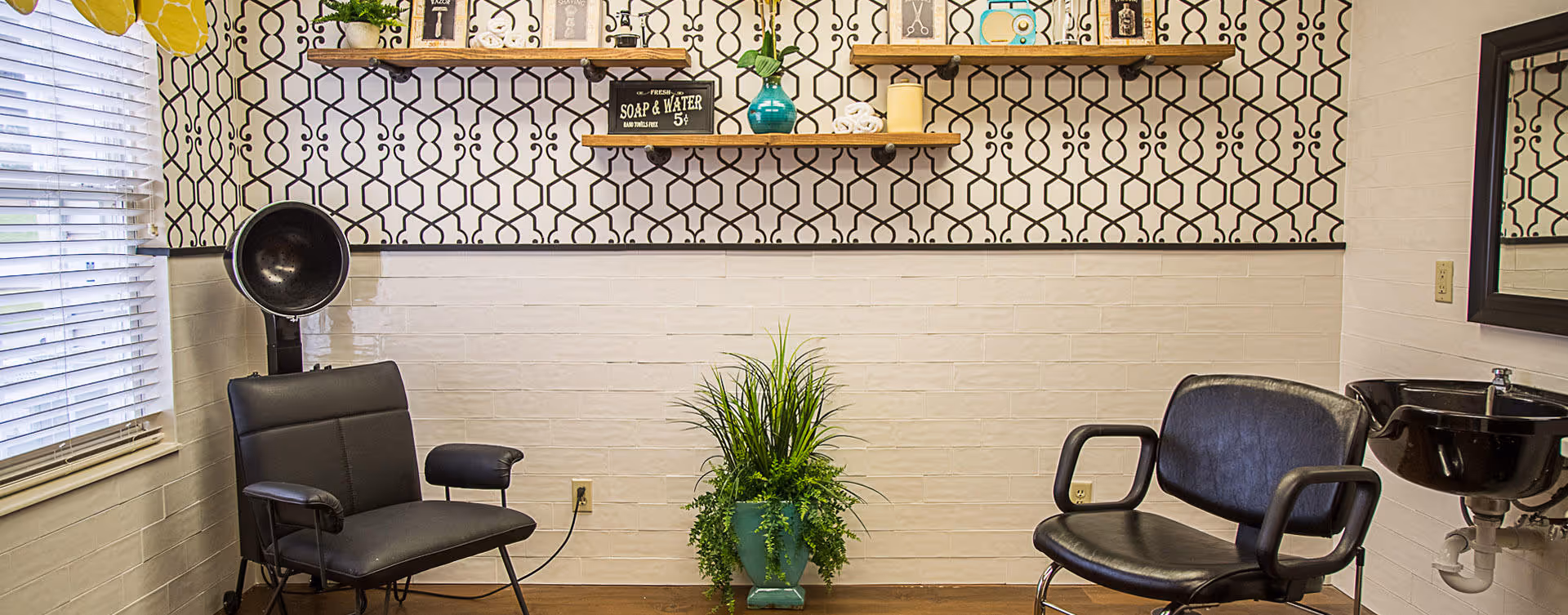 Interior of a salon or hair styling area with two black salon chairs, a black hair dryer, a black sink with a mirror above it, a green plant in a blue pot on the floor, and a wall decorated with patterned wallpaper and shelves holding various decorative items.