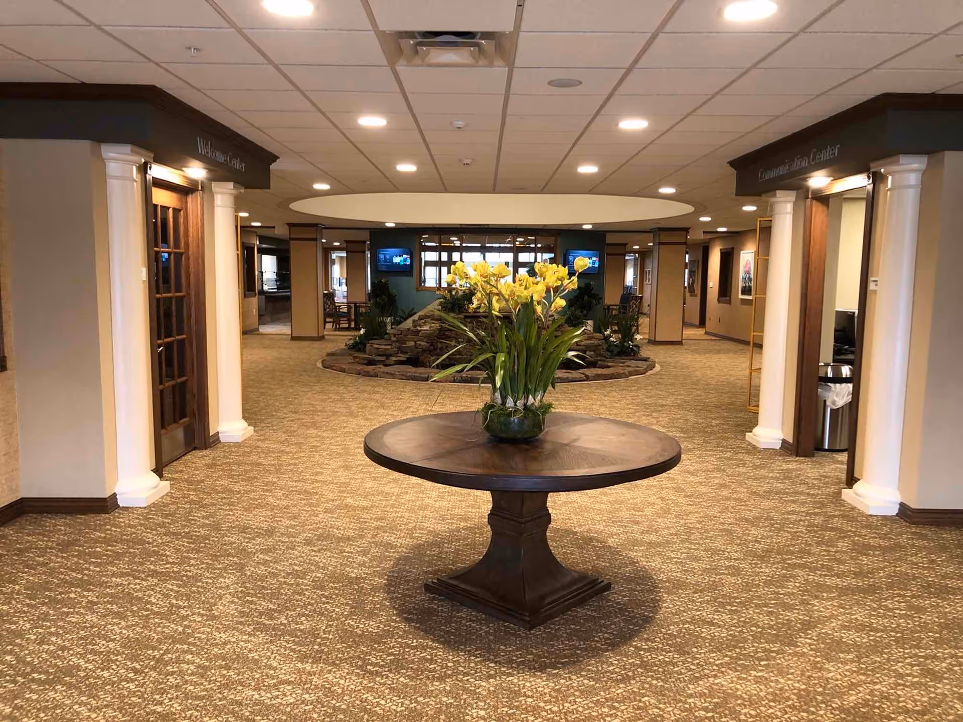 Interior view of a spacious hallway in a retirement community with a round wooden table in the center holding a vase of yellow flowers. The hallway has carpeted floors, white columns, and doors labeled 'Wellness Center' and 'Examination Center'. There is a rock garden feature with plants and two television screens mounted on the far wall.