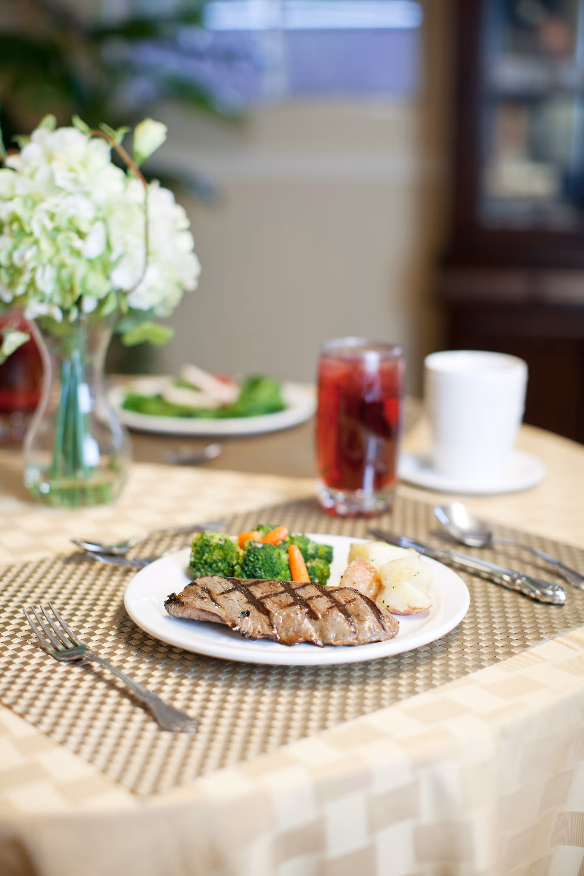 A plated grilled steak with broccoli, carrots and potatoes on a table with a drink and a vase of flowers in the background.