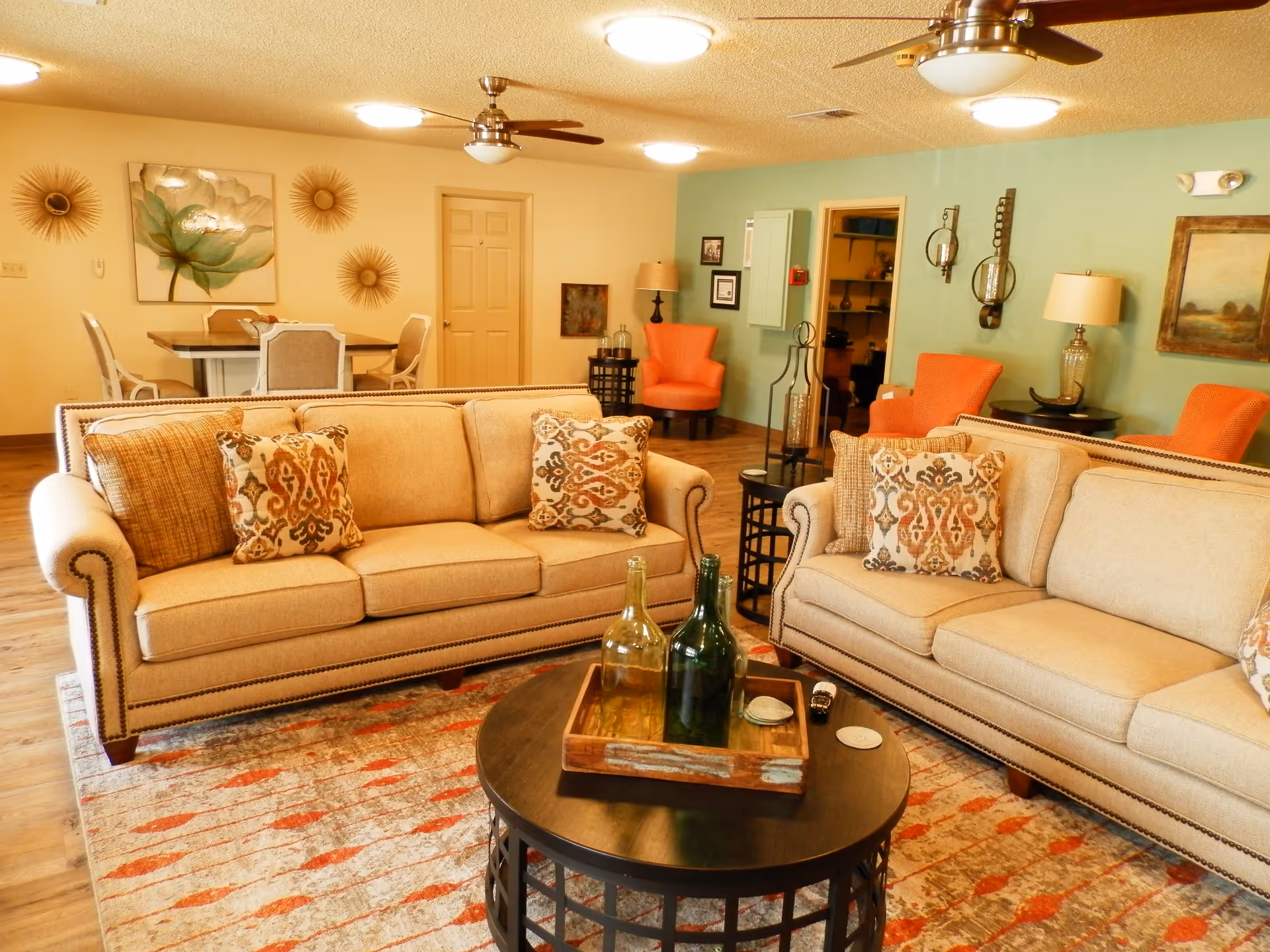 A cozy living room with two beige sofas adorned with patterned cushions, a round black coffee table with decorative bottles on a wooden tray, and a colorful area rug. The background features a dining table with chairs, orange armchairs, wall art, and warm lighting fixtures including ceiling fans with lights.