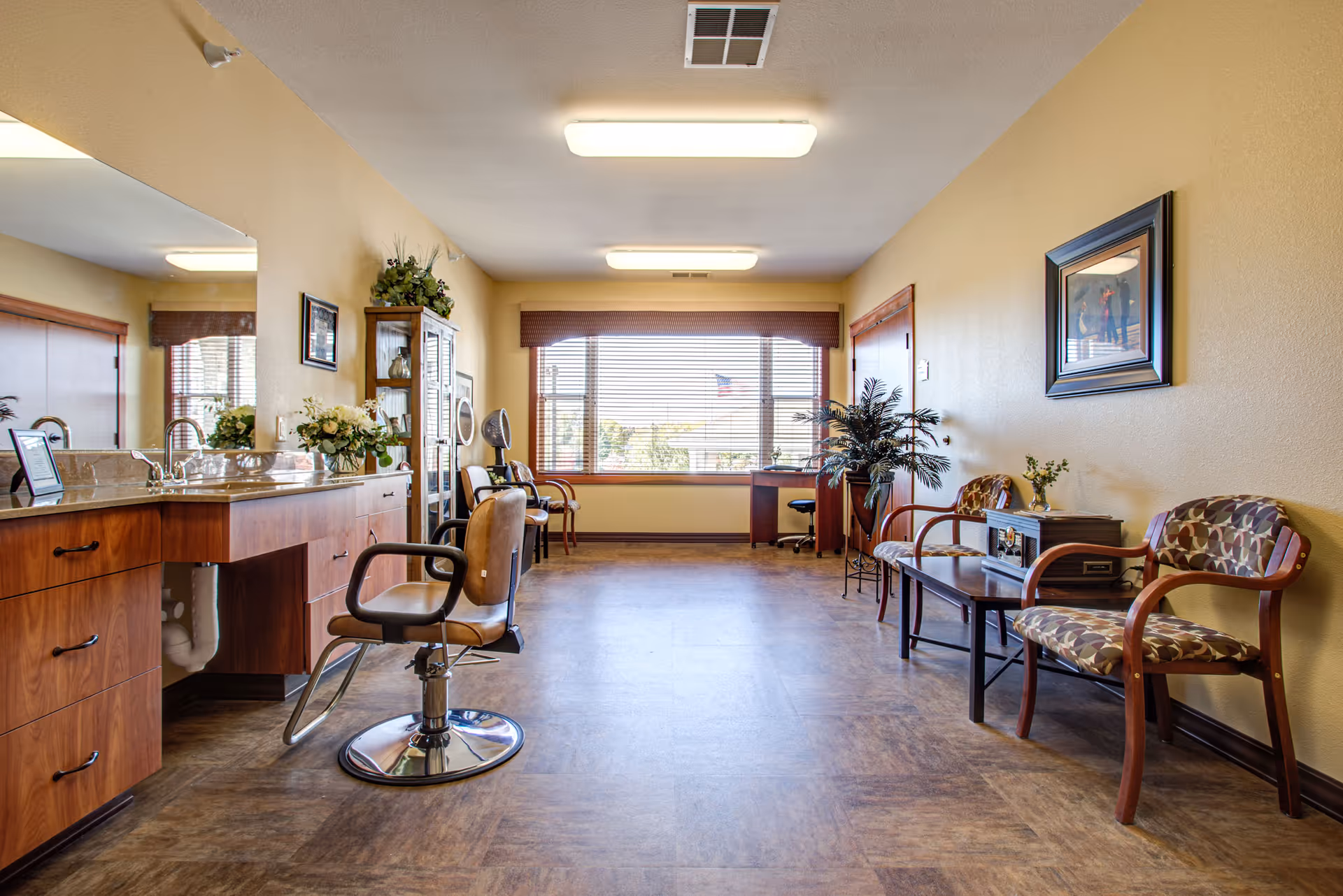 A bright and spacious room in a senior living facility with a large window at the far end letting in natural light. The room features a salon-style chair in front of a counter with a sink and mirror on the left side. On the right side, there are two patterned armchairs with a small table between them, decorated with a plant and a framed picture on the wall above. The floor has a wood-like finish, and the walls are painted a warm beige color.