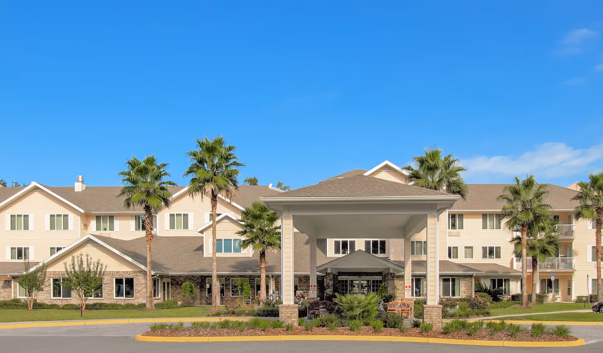 Front exterior view of Marion Woods by Sky Active Living, a multi-story senior living facility with beige siding and stone accents, surrounded by palm trees and landscaping under a clear blue sky.