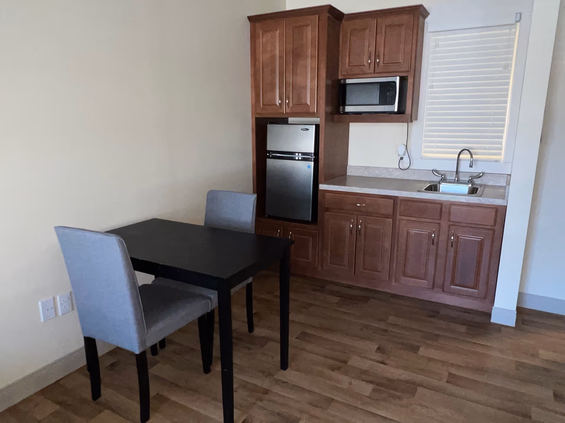 Small kitchenette with wooden cabinets, a sink, microwave, mini fridge and a black dining table with two gray chairs.