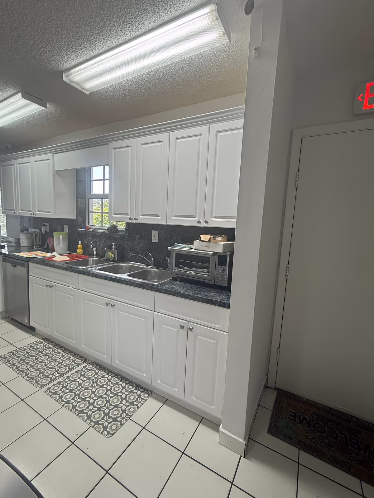 A clean kitchen with white cabinets and drawers, a black countertop, a double sink, a toaster oven, and various kitchen items on the counter. The floor has white tiles with two patterned rugs in front of the sink area. There is a window above the sink letting in natural light, and fluorescent ceiling lights illuminate the space. A door with an exit sign above it is visible on the right side.