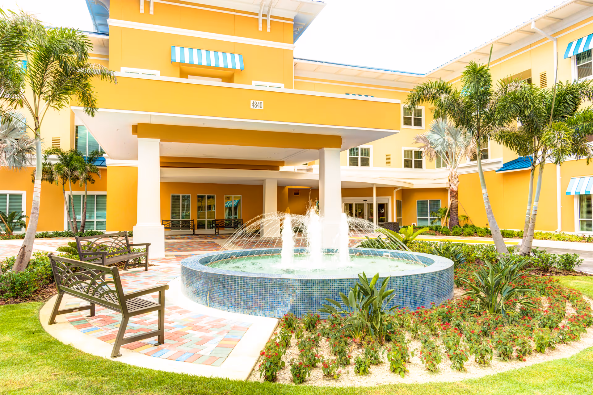 Outdoor courtyard area of a senior living facility with a circular water fountain surrounded by plants and flowers. There are benches on a colorful tiled pathway, palm trees, and a yellow building with blue and white striped awnings in the background.
