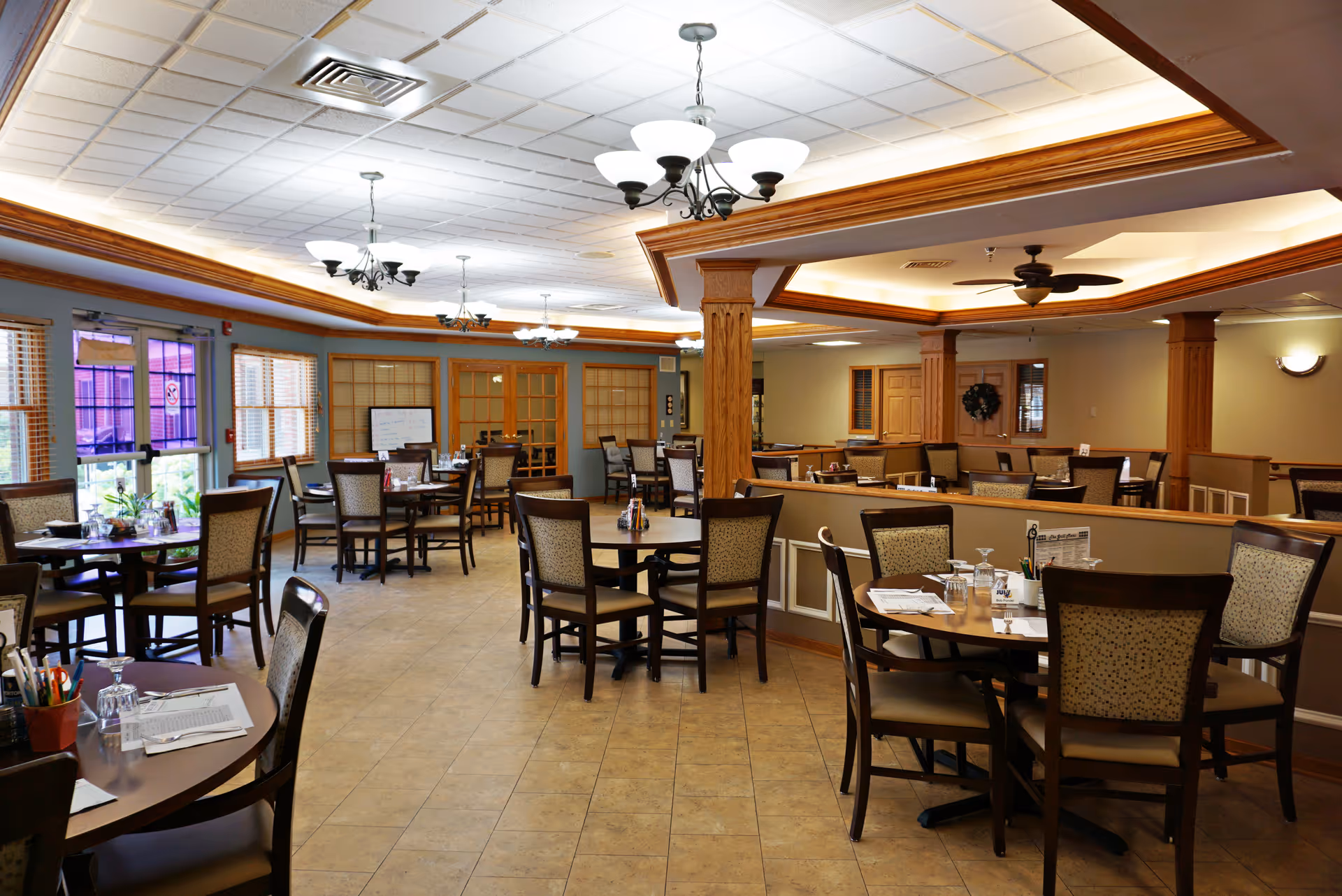 A spacious dining room in an assisted living facility with multiple round tables and chairs arranged neatly. The room features tiled flooring, wooden trim, ceiling fans, and chandeliers. Windows and glass doors allow natural light to enter the room.
