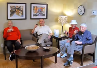 Four elderly people sitting in a living room area, each holding a glass and smiling. The room has a coffee table with decorative bowls, two framed pictures on the wall, a table lamp, a clock, and a thermostat on the wall.
