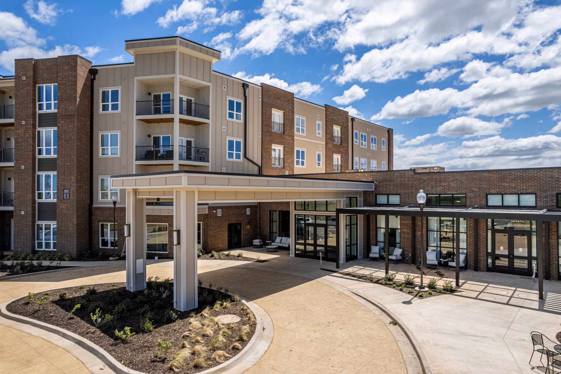 Exterior view of a modern senior living facility building with a covered entrance, multiple floors with balconies, large windows, and a landscaped driveway under a partly cloudy sky.