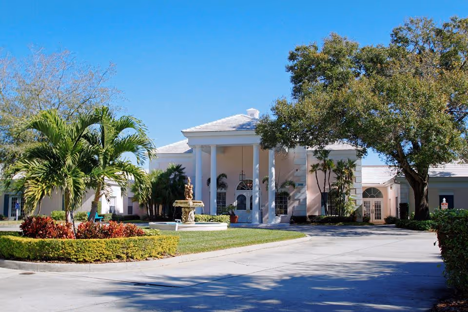 Front exterior view of a senior living facility with a circular driveway, a fountain in the center, palm trees, and a large tree on the right under a clear blue sky.