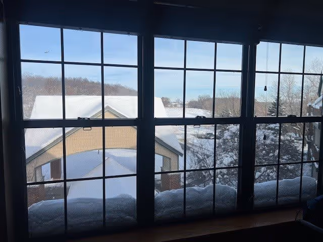 View through large multi-pane windows showing a snowy outdoor scene with rooftops covered in snow and trees in the background under a clear blue sky.