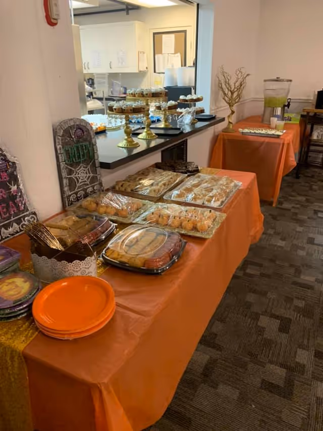 A buffet table covered with an orange tablecloth displaying various baked goods and snacks, including cookies, cupcakes on gold stands, and other treats. There are Halloween-themed decorations such as tombstone signs and orange plates. In the background, there is a beverage dispenser with a green drink and a small table with additional items.