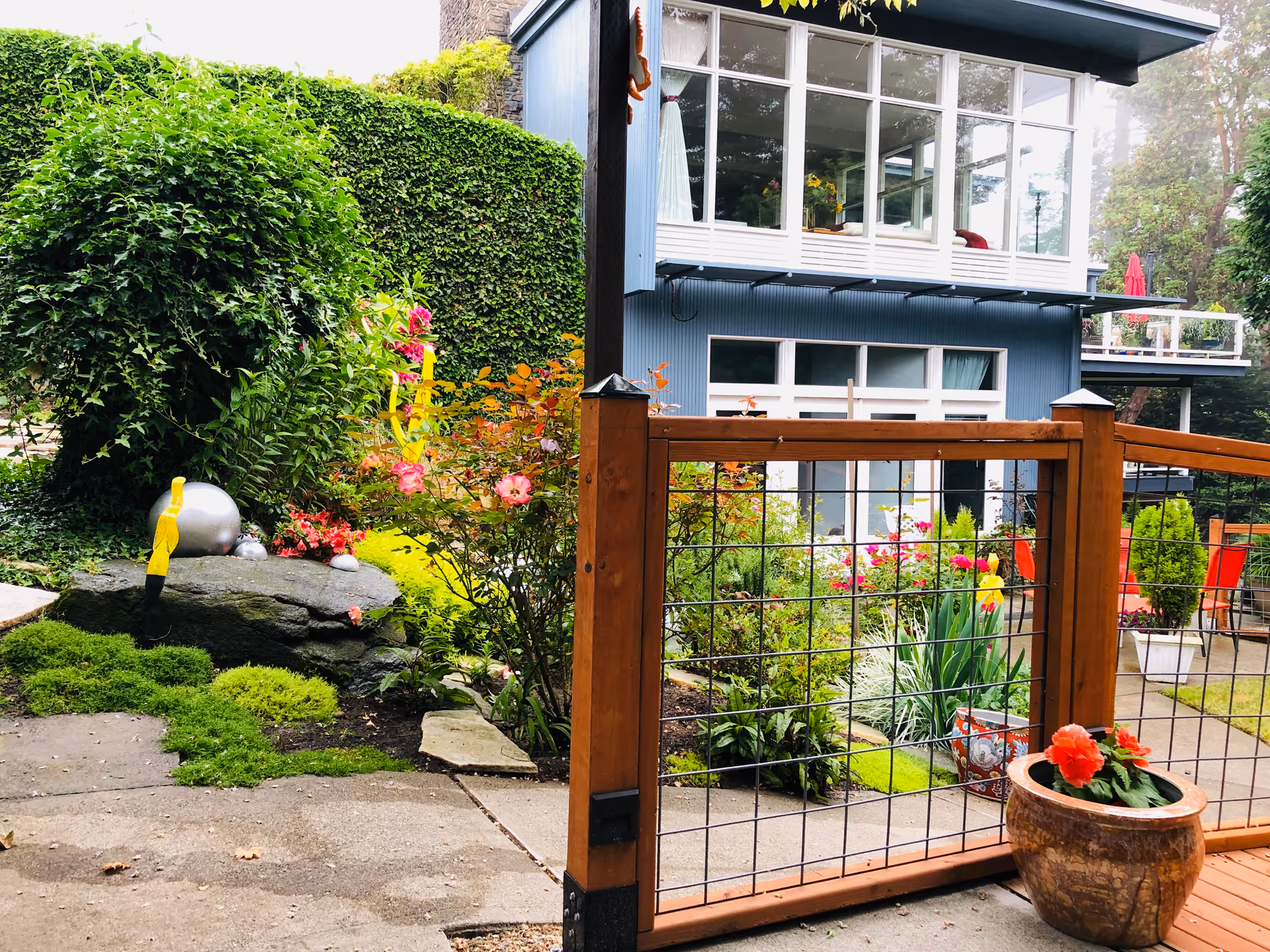 Landscaped outdoor garden and patio with a wooden wire fence and potted flowers in front of a two-story blue building with large windows.