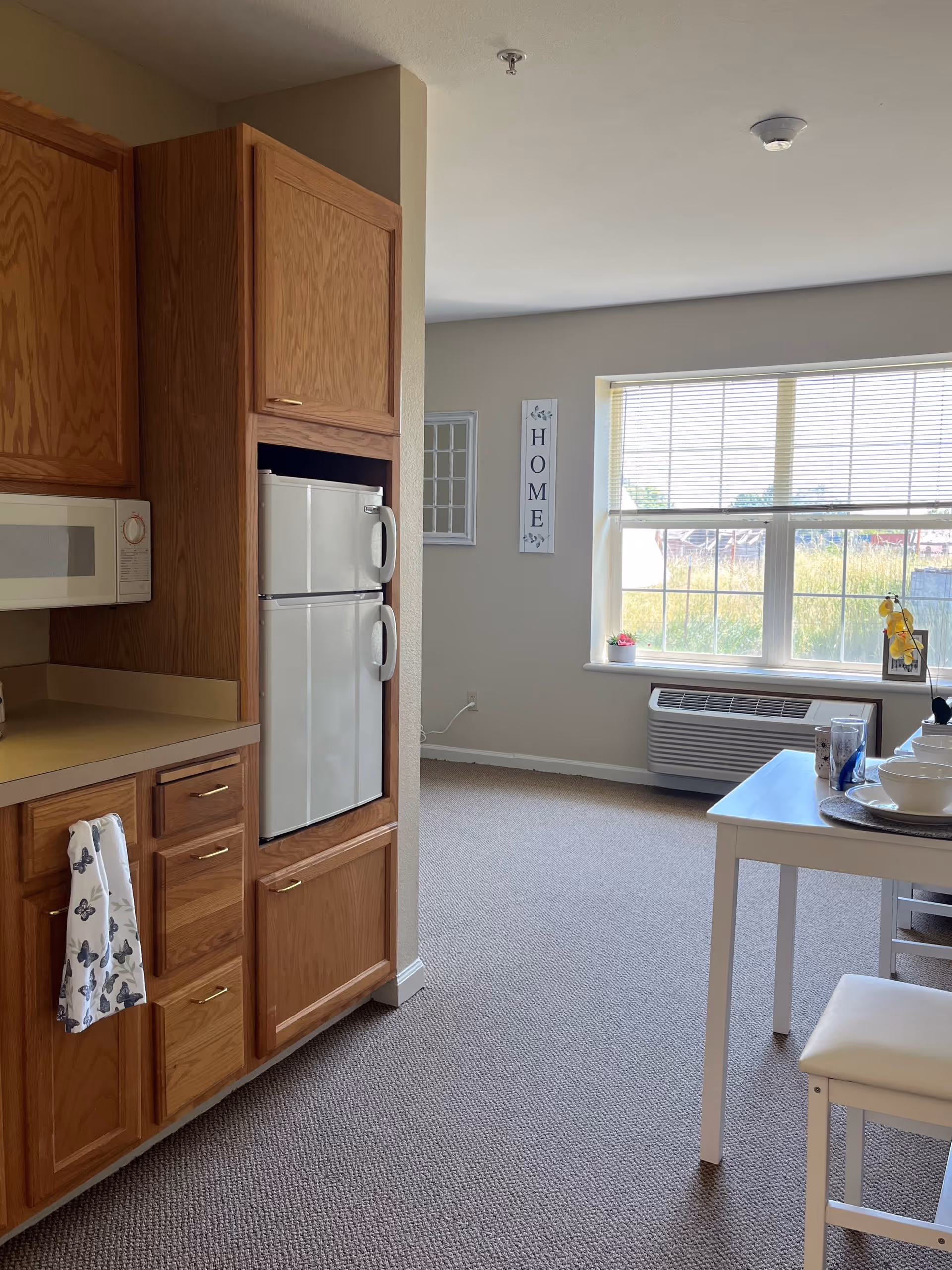 Interior view of a senior living facility apartment showing a small kitchen area with wooden cabinets, a white microwave, and a white mini refrigerator. Adjacent to the kitchen is a dining table set with white dishes and a chair. A large window with blinds lets in natural light, and a wall decoration with the word 'HOME' is visible. The floor is carpeted and there is an air conditioning unit below the window.