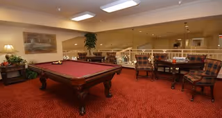 Interior view of a recreational room with a red carpet, a pool table with balls arranged, a small table with four plaid upholstered chairs, a lamp on a side table, a large mirror on the wall, and a potted plant in the corner.