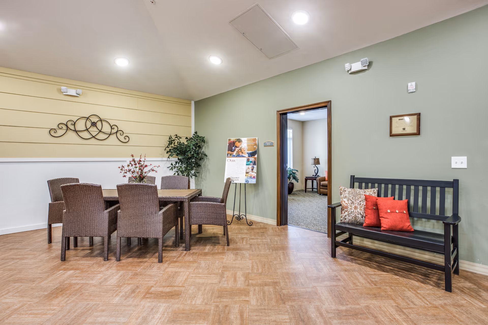 Common area with a wicker dining table and chairs, a potted plant, and a black bench with red pillows against a green wall.