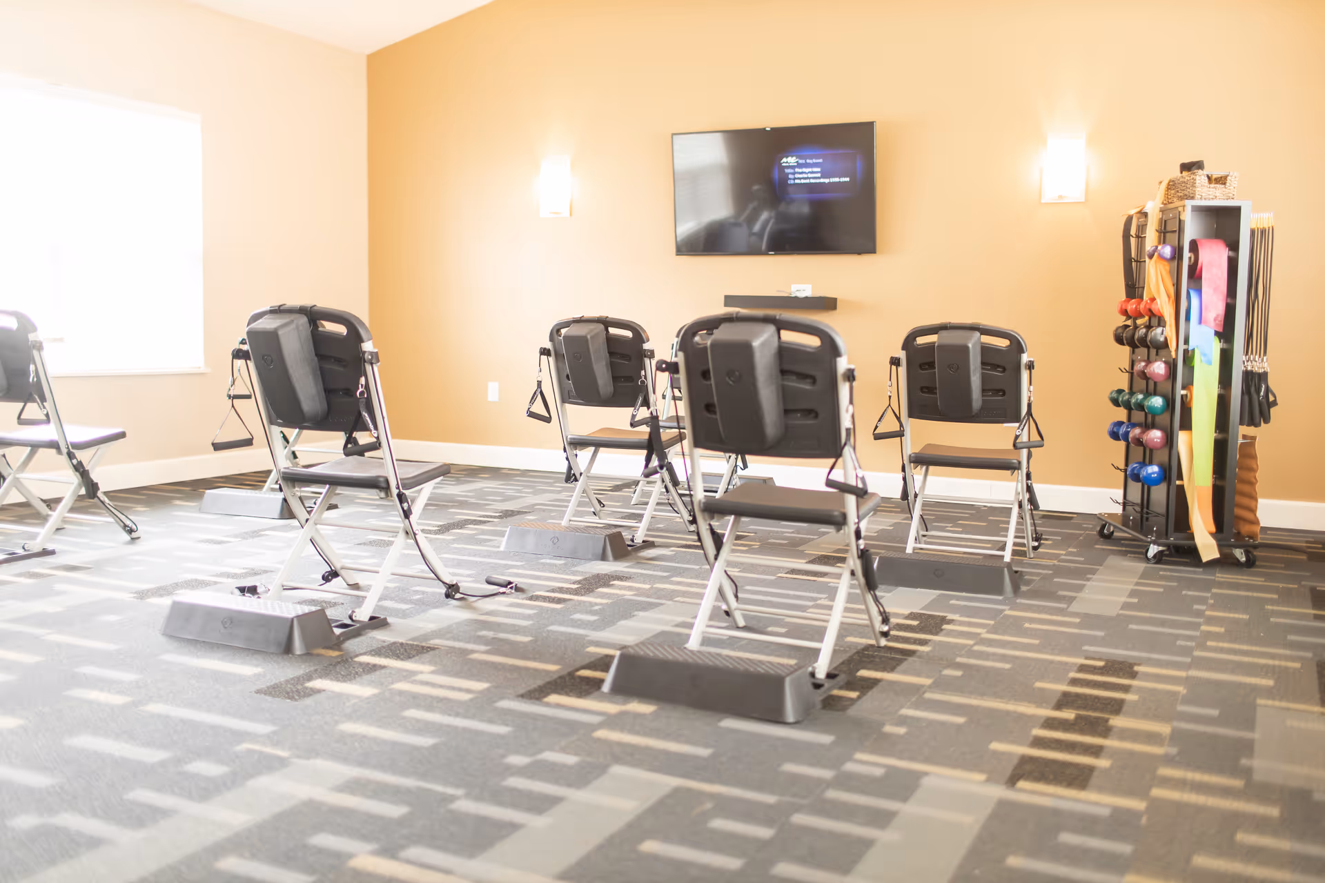 A bright exercise room with several black folding chairs equipped with resistance bands and foot platforms arranged in rows. A rack holding colorful dumbbells and exercise bands is visible on the right side of the room. A flat-screen TV is mounted on a beige wall at the front of the room.