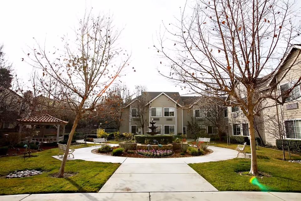 Outdoor courtyard area at Aegis Gardens Fremont featuring a circular walkway surrounding a central fountain with flowers and decorative elements. There are benches along the path and leafless trees, with a gazebo to the left and residential buildings in the background.