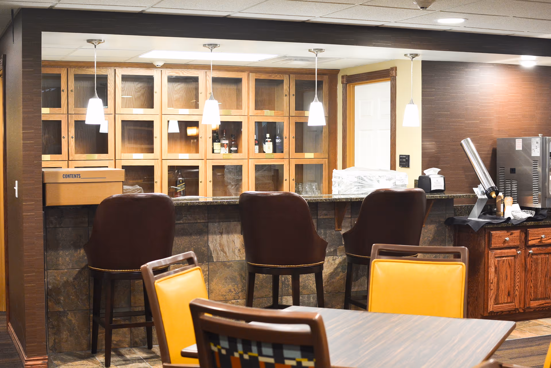 Interior view of a bar area in a retirement community with three brown leather bar stools in front of a stone and wood counter. Behind the counter are wooden cabinets with glass doors containing bottles and glassware. Pendant lights hang from the ceiling above the counter. In the foreground, there is a table with chairs, one chair having a yellow cushion.