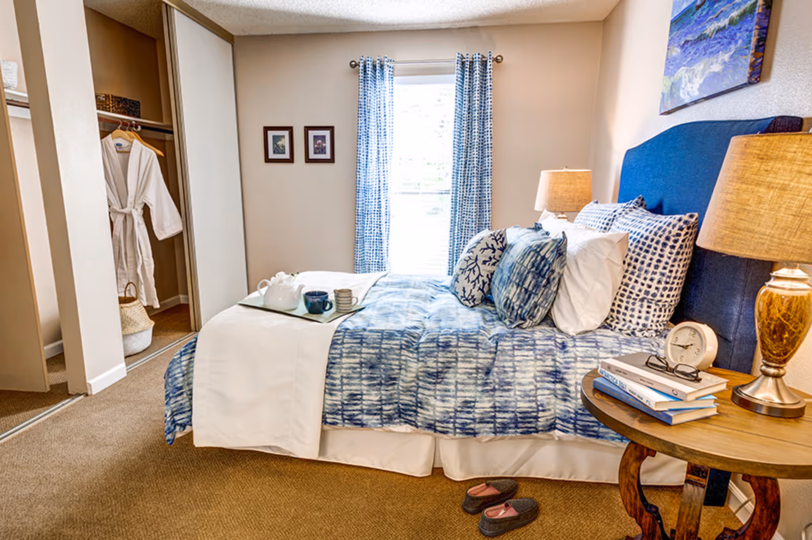 A cozy bedroom with a bed dressed in blue and white patterned bedding and multiple pillows. A tray with a teapot and cups is placed on the bed. To the right of the bed is a wooden nightstand with a lamp, an alarm clock, a pair of glasses, and a stack of books. A pair of slippers is on the carpeted floor near the bed. The room has a window with blue and white curtains, two framed pictures on the wall, and an open closet with a white robe hanging inside.