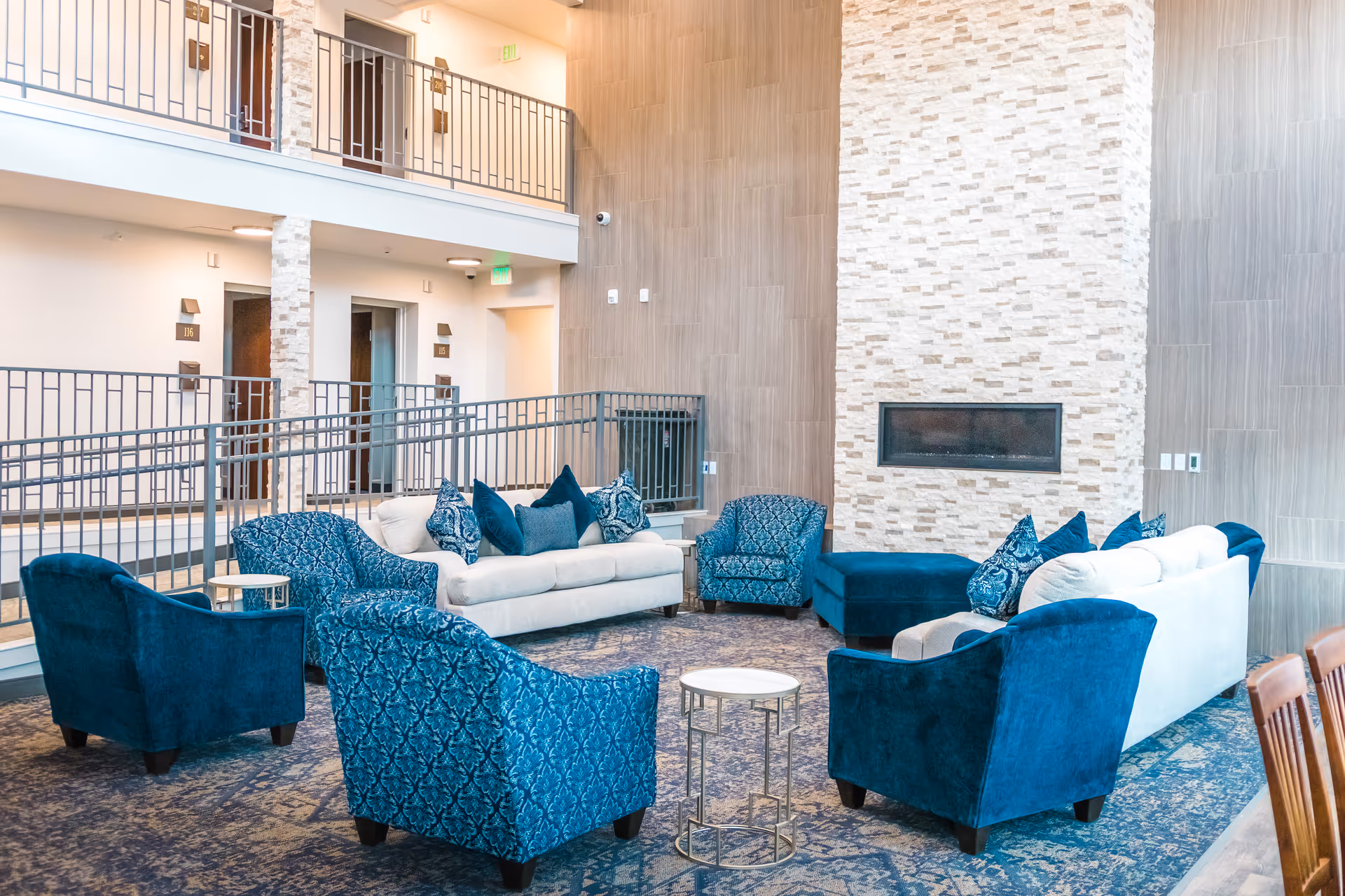 A spacious and modern common area in an assisted living facility featuring blue and white upholstered sofas and armchairs arranged around a small round table. The room has a tall stone fireplace, light wood paneling on the walls, and a patterned carpet. There is a second-floor balcony with railings and doors to individual rooms visible in the background.