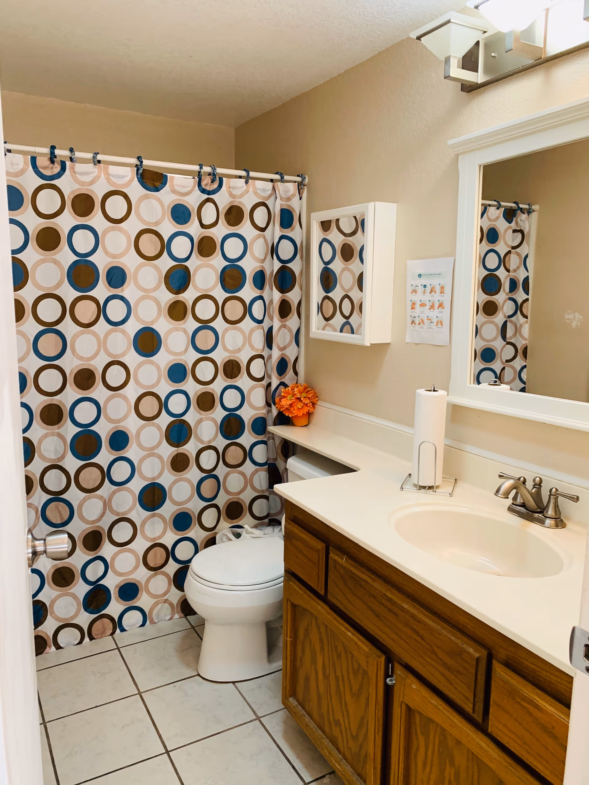 A bathroom with a white toilet, a wooden vanity with a white countertop and sink, a mirror above the sink, a wall-mounted medicine cabinet, a colorful shower curtain with blue, brown, and beige circles, tiled floor, and a small orange flower arrangement on the countertop.