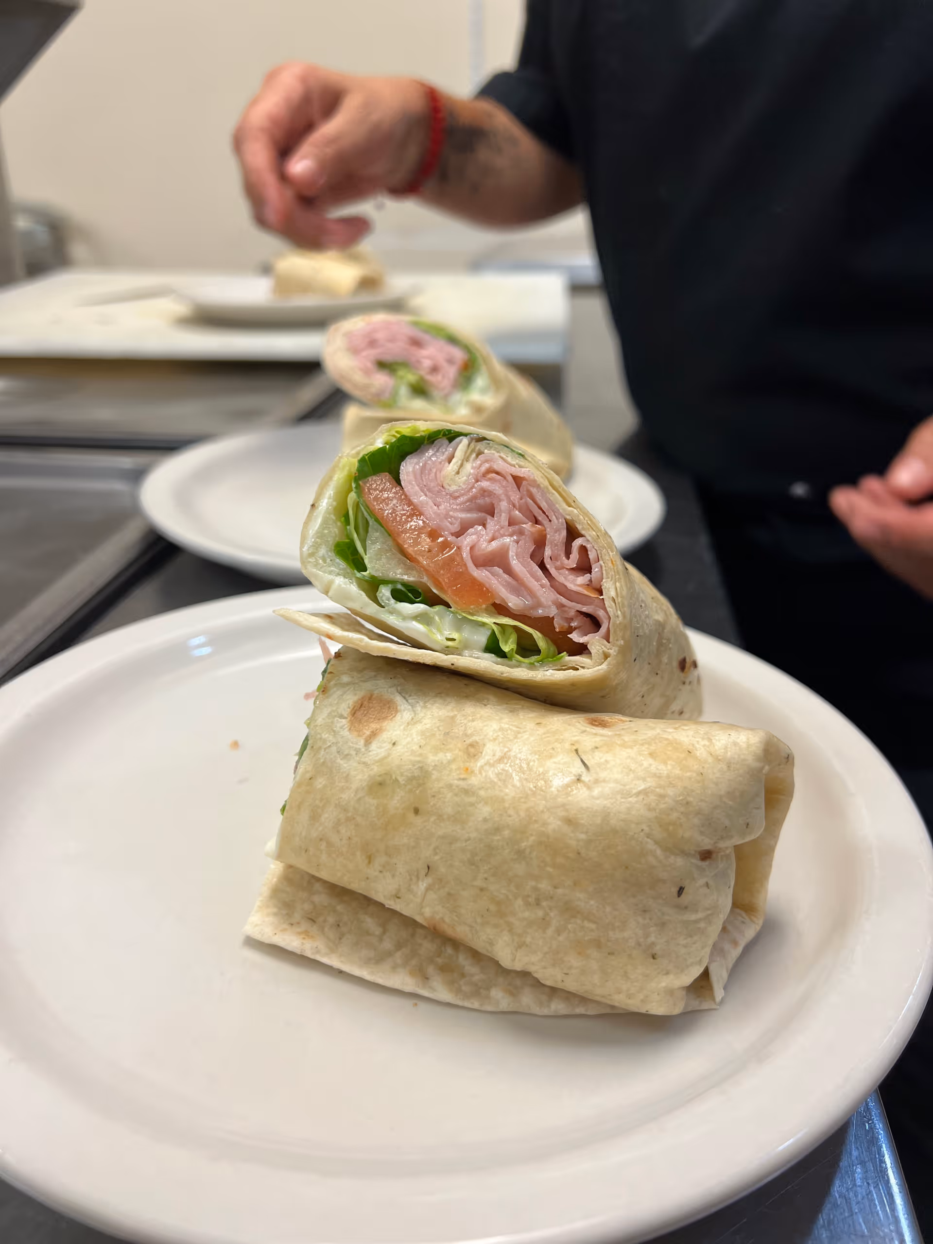Halved ham-and-lettuce tortilla wraps stacked on a white plate with a person preparing more wraps in a kitchen background.