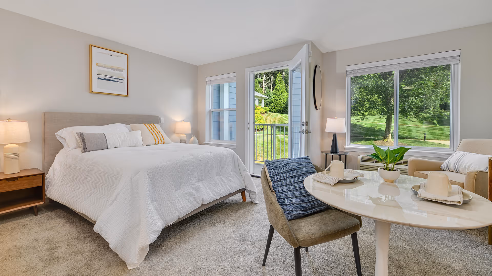 A bright and cozy bedroom with a large bed covered in white bedding, two bedside tables with lamps, a round table set with two chairs, a potted plant, and large windows showing a green outdoor view.