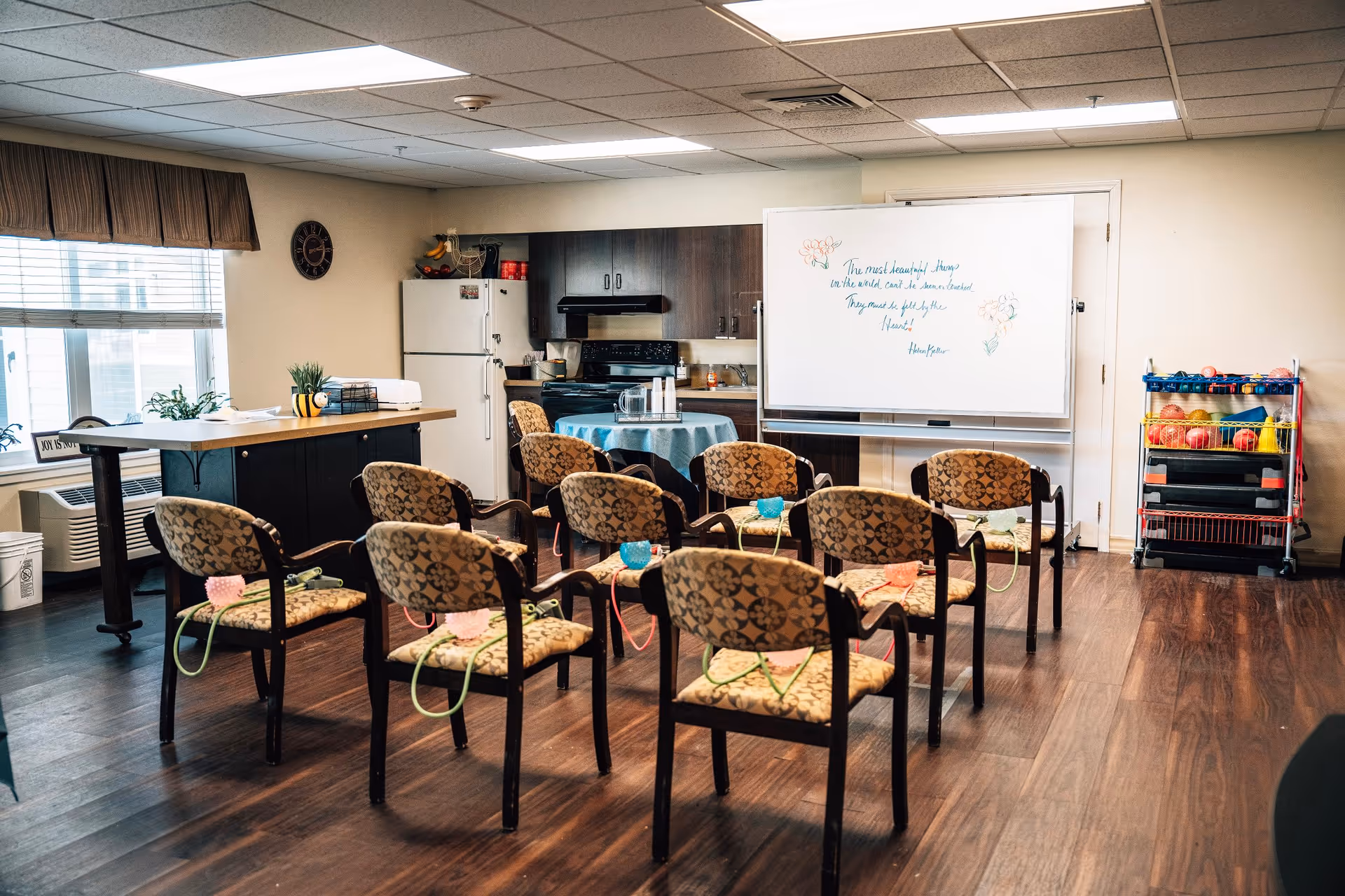 A room with several patterned chairs arranged in rows facing a whiteboard with a handwritten quote. Behind the chairs is a small kitchen area with a refrigerator, stove, and cabinets. There is a table covered with a blue cloth and a rack with colorful balls on the right side. The room has wooden flooring and a window with blinds and a valance.