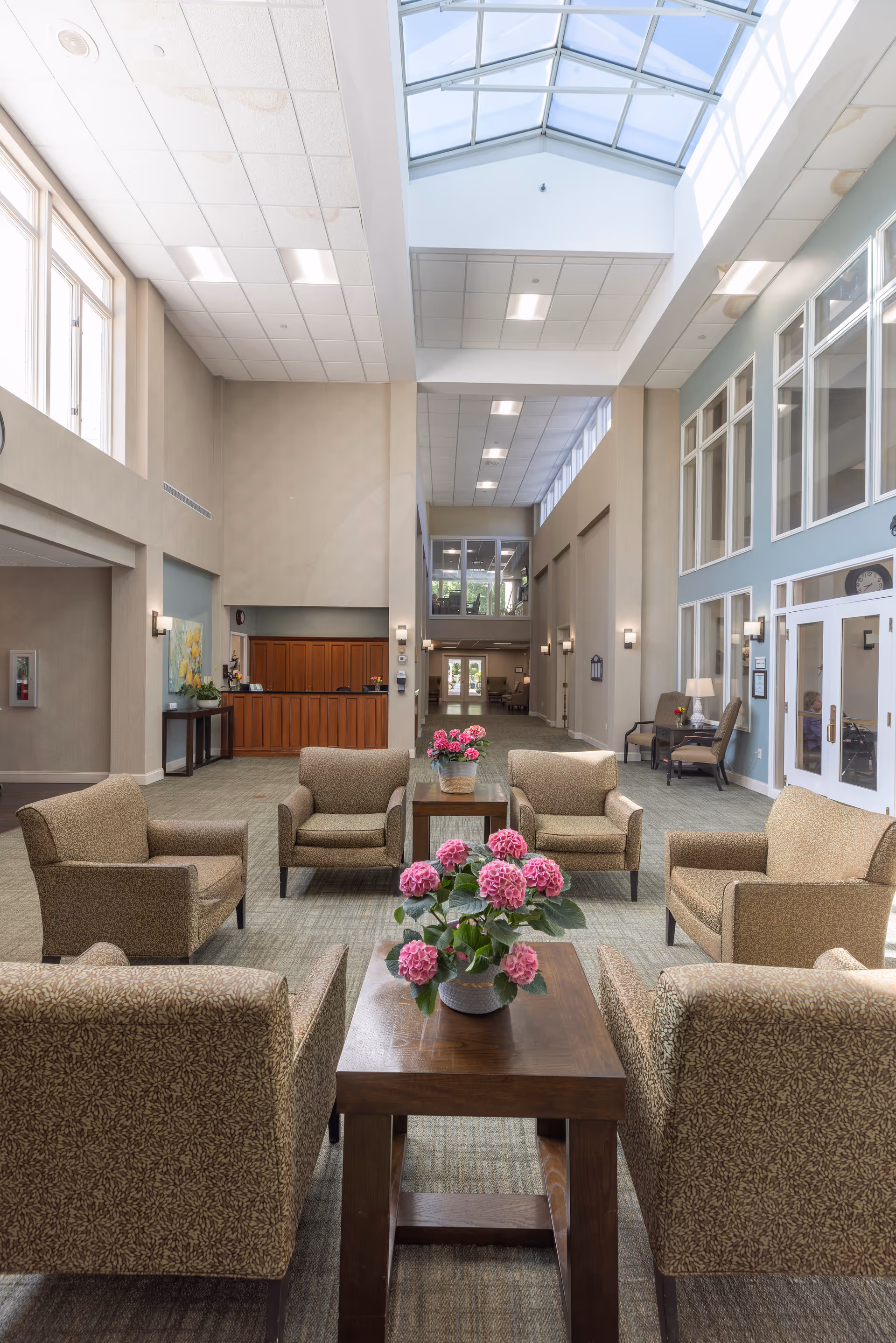 A spacious and well-lit common area in an assisted living facility featuring a high ceiling with a large skylight. The room has beige walls and carpeted floors. There are six patterned armchairs arranged around two wooden tables, each table holding a pot of pink flowers. In the background, there is a reception desk and large windows allowing natural light to enter.