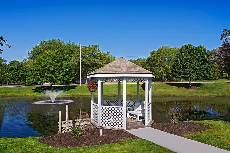 A white wooden gazebo with a shingled roof situated next to a pond with a water fountain. The gazebo has lattice panels and hanging flower baskets, surrounded by green grass and trees under a clear blue sky.
